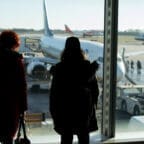 Two people watch through an airport window as passengers board a plane on the tarmac
