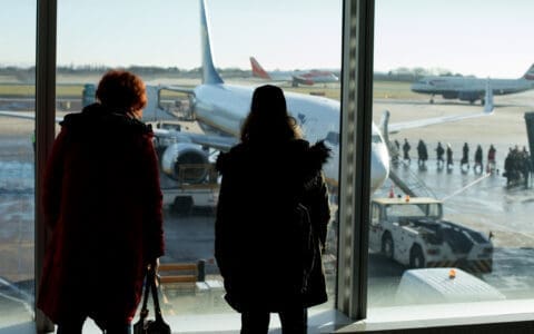Two people watch through an airport window as passengers board a plane on the tarmac