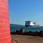 A red lighthouse base beside a harbor, with a ferry sailing past on the sea toward the coastline
