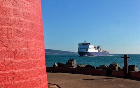 A red lighthouse base beside a harbor, with a ferry sailing past on the sea toward the coastline